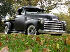 an old black truck is parked on the grass in front of some trees and leaves