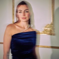 a woman in a blue dress standing next to a gold framed wall with a diamond necklace on it