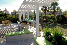 a covered patio area with green cushions and white pergolated roof, surrounded by palm trees