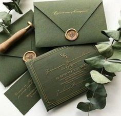 some green envelopes with wax stamp on them next to eucalyptus leaves and greenery