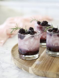 two glasses filled with ice and blackberries on a wooden tray next to someone's hand