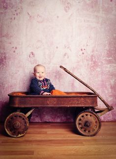 a baby sitting in a wooden wagon with wheels on the floor next to a pink wall