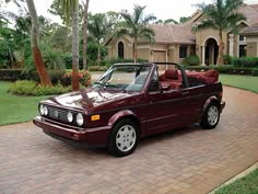 a maroon convertible car parked in front of a house with palm trees and bushes behind it