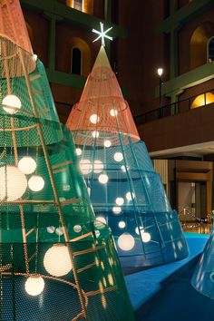 three brightly colored christmas trees with lights on them in an indoor building lobby, one is lit up and the other is made out of mesh