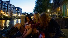 three young women sitting on the edge of a river at night looking at their cell phones