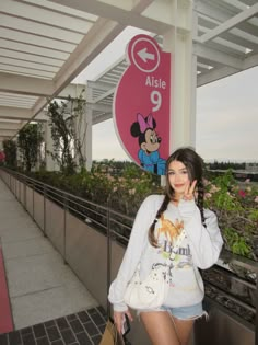 a woman standing next to a sign with minnie mouse on it's back and holding a handbag