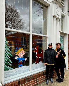 two people standing in front of a store window with snow on the windows and christmas decorations behind them