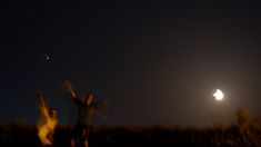 two people are standing in the grass at night with their arms up and one person is reaching for an object