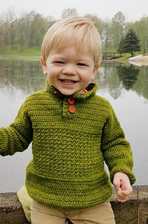 a little boy standing next to a body of water wearing a green sweater and khaki pants