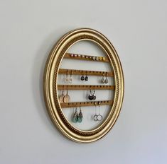 an oval gold frame holds earrings and earring hooks on a white wall behind it