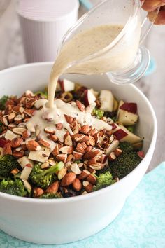 a bowl filled with broccoli, apples, nuts and dressing being poured into it