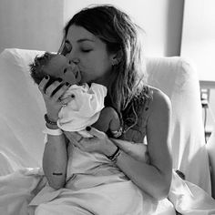 a woman holding a baby in her lap while sitting on a bed and kissing it's face