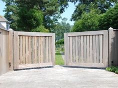 two wooden gates are open in front of a brick walkway and tree - lined yard