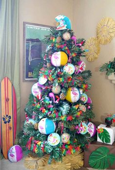 a colorful christmas tree decorated with beach balls and palm leaves, sits in front of a surfboard