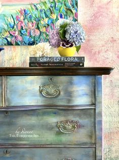 an old dresser with flowers and a book on it's top sits in front of a floral painting