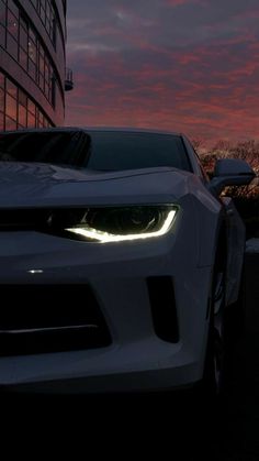 a white car parked in front of a tall building at night with the sun going down