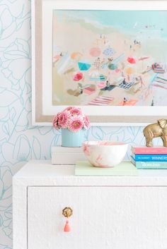 a white dresser topped with pink flowers next to a wallpapered wall and painting