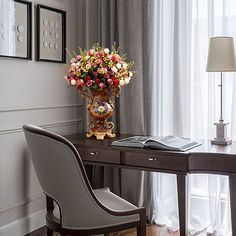 a desk with flowers in a vase on top of it next to a chair and lamp