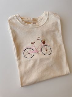 a t - shirt with a pink bicycle embroidered on the front and back, sitting on a white surface