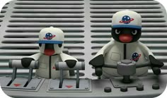 two toy figures sitting on top of a metal object with holes in the middle and one is wearing a baseball cap