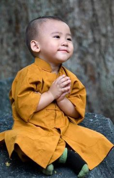 a little boy sitting on top of a rock with his hands folded in front of him