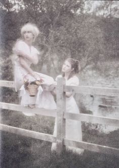 two women in white dresses standing on a wooden fence next to a river and trees