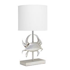 a table lamp with a white shade on it and a silver crab figure underneath the light