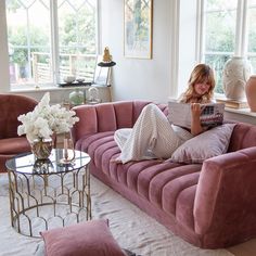 a woman sitting on a pink couch reading a book in a living room with large windows