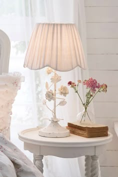 a white table with flowers and a book on it in front of a lamp that is turned on