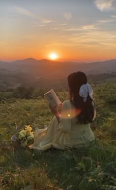 a woman sitting on top of a lush green hillside reading a book at sunset with the sun setting behind her