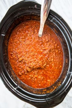 a spoon in a slow cooker filled with chili and meat sauce on a marble counter top