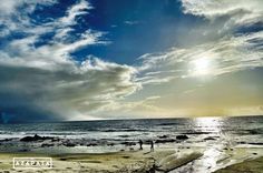 two people are walking on the beach near the ocean under a cloudy sky with sunbeams