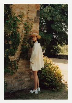 a woman standing next to a brick wall wearing a white dress and hat with ivy growing on it