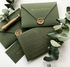 some green envelopes with wax stamp on them next to eucalyptus leaves and greenery