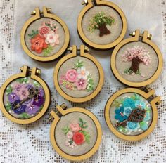 six embroidered flowers are in wooden frames on a white tablecloth with lace and beads