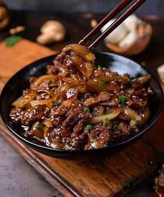 a black bowl filled with beef and onions on top of a wooden cutting board next to chopsticks