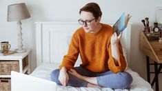 a woman is sitting on her bed reading a book and looking at the laptop screen