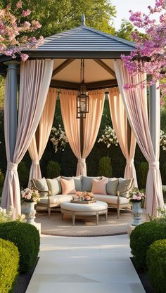 a gazebo with curtains and couches in the middle of a garden area surrounded by flowers