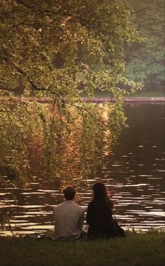 two people are sitting on the grass by the water and looking at the trees in the distance