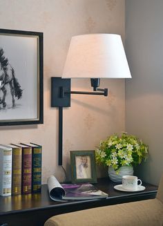 a lamp on a wall next to a table with books and a vase filled with flowers