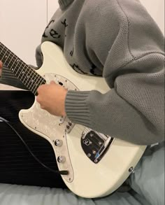 a person sitting on a bed playing an electric guitar while wearing a gray sweater and black pants