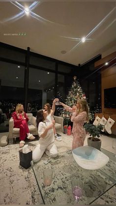 three women are sitting on the floor in front of a christmas tree and one woman is reaching for something