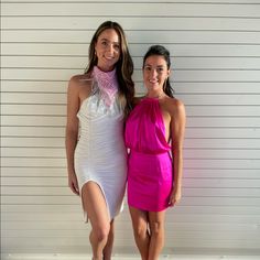 two women standing next to each other in front of a white wall and one is wearing a pink dress