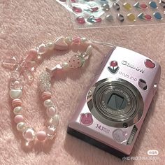 a camera and some beads on a pink blanket next to a necklace that says hello kitty