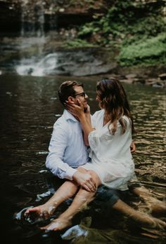 a man and woman are sitting in the water near a waterfall, holding each other