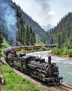 an old fashioned steam train traveling down the tracks near a river and mountains in the background