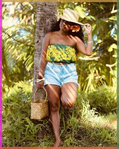 a woman in a yellow and green top leaning against a tree holding a straw bag