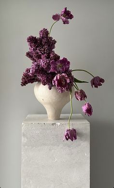 a vase with purple flowers in it sitting on top of a white block and gray background