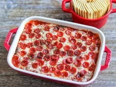 a red casserole dish filled with pepperoni and cheese next to a bowl of crackers