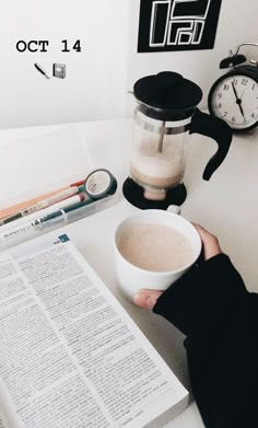 a person holding a coffee cup in front of an open book and clock on a desk